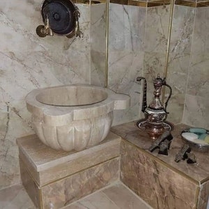 Puede incluir: Un lavabo de mármol blanco con un diseño decorativo se asienta sobre un pedestal de mármol a juego. El lavabo se encuentra en un baño con azulejos de mármol beige y una tetera de cobre con un diseño decorativo en una bandeja a juego.