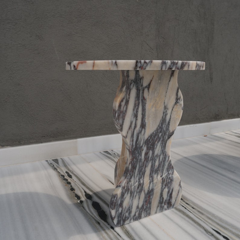 May include: A round, white marble table with a unique, organic-shaped base. The table has a swirling pattern of grey and pink veins.