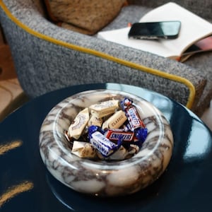 May include: A white marble bowl filled with assorted chocolate candy bars, including Snickers, Milky Way, and 3 Musketeers. The bowl is sitting on a dark blue table.