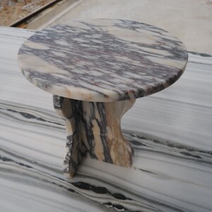 May include: A small, round, white marble table with a unique, swirling, purple and brown pattern. The table has a simple, curved, pedestal base.
