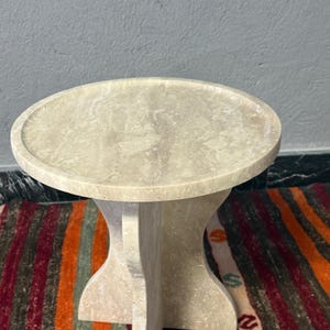 May include: A beige stone side table with a round top and a unique pedestal base. The table is approximately 50 cm in diameter and 50 cm tall. The table is set on a colorful striped rug.