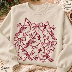 May include: A sand-coloured crewneck sweatshirt featuring a dark red graphic print of four cardinal birds, three decorative bows, and several small flowers arranged in a pattern across the centre of the garment.
