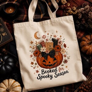 May include: A cream-colored canvas tote bag featuring a graphic print of an orange jack-o'-lantern topped with books and flowers. A black bow sits on the pumpkin. The text reads "Booked for Spooky Season" in black script below the illustration.