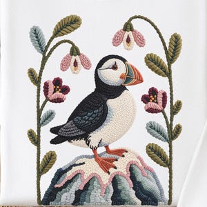 May include: White sweatshirt featuring a colorful embroidered puffin bird design. The puffin is perched on a rock with flowers and foliage. The design includes shades of black, white, blue, green, pink, and orange. The sweatshirt has long sleeves and a crew neck.