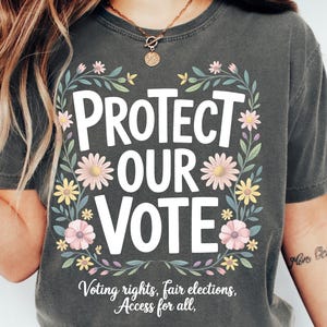 May include: Dark gray t-shirt with the words "PROTECT OUR VOTE" in white, surrounded by a floral wreath. The shirt also includes the text "Voting rights, fair elections, Access for all."