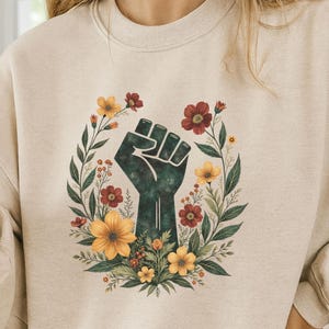 May include: Beige sweatshirt featuring a floral wreath design with a raised fist graphic. The wreath includes yellow, red, and green elements. The sweatshirt has long sleeves and a crew neck.