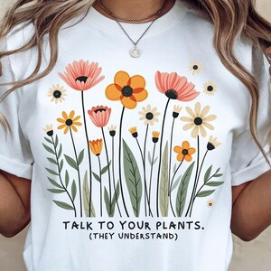 May include: White t-shirt featuring a colorful floral design with orange, pink, and yellow flowers, green leaves, and the text "TALK TO YOUR PLANTS. (THEY UNDERSTAND)". The shirt has a crew neck and short sleeves.