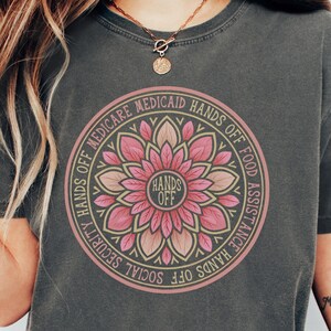 May include: A dark gray t-shirt with a pink and gold mandala design. The mandala features the words "Hands Off" in the center and a circle of text around the outside that reads "Hands Off Social Security Hands Off Food Assistance Medicare Medicaid Hands Off".