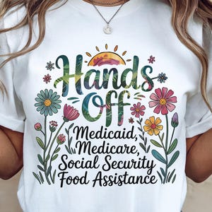 以下が含まれることがあります： 花柄のデザインと「Hands Off Medicaid, Medicare, Social Security, Food Assistance」というテキストが書かれた白いTシャツ。