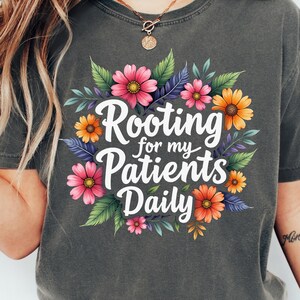 May include: Dark gray t-shirt with a floral design and the text "Rooting for my Patients Daily." The design features pink, orange, and yellow flowers with green and purple leaves. A gold necklace is visible.