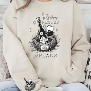 May include: A beige sweatshirt with a black graphic design of a wine bottle and glass in a nest. The text on the sweatshirt reads "This Empty Nester est 2025 has PLANS".