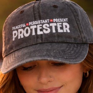 May include: A grey baseball cap with the word "PROTESTER" in white block letters. Above the word are the words "PEACEFUL * PERSISTANT * PRESENT". The cap is worn by a person with long, wavy auburn hair.