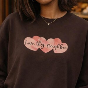 May include: A dark brown sweatshirt featuring the phrase "Love thy neighbor" in a watercolour design. The text is above three pink hearts. The sweatshirt is plain and has long sleeves.