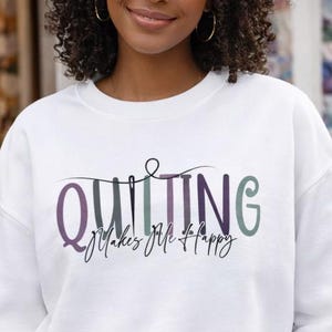 May include: A white crewneck sweatshirt featuring the text "QUILTING" in large, multi-colored letters with a needle and thread graphic, and the script text "Makes Me Happy" underneath. The garment has long sleeves and a ribbed collar.