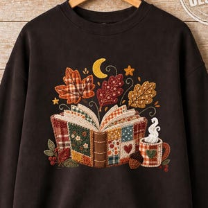 Pode incluir: Uma camisola de gola redonda castanho chocolate escuro com um bordado central de um livro aberto estilo patchwork, folhas de outono, uma lua crescente, estrelas e uma caneca a fumegar, com o texto ORIGINAL DESIGNER no canto superior direito.