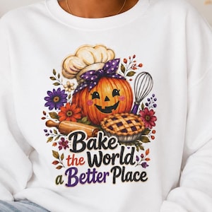 May include: A white crewneck sweatshirt featuring a colourful graphic of a jack-o'-lantern wearing a chef's hat and a purple polka-dot bow. The design includes a rolling pin, a whisk, a pie, and flowers, with the text "Bake the World a Better Place."