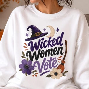 May include: A white crewneck sweatshirt featuring a graphic print with the text Wicked Women Vote in purple and black script. The design includes a purple witch hat, a crescent moon, small stars, a broomstick, and purple and white flowers.