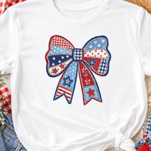 May include: A white short-sleeved t-shirt featuring a large, central graphic of a decorative bow. The bow is composed of various patchwork-style patterns in red, white, and blue, including stars, stripes, gingham checks, and small floral motifs.