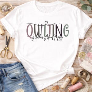 Peut inclure: T-shirt blanc avec l'inscription "Quilting Makes Me Happy" dans une police décorative. Le mot "Quilting" est dans des tons violets et verts, avec un motif d'aiguille et de fil. Le t-shirt est présenté avec des fournitures de couture.