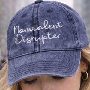 May include: A navy blue baseball cap featuring the embroidered words "Nonviolent Disrupter" in white script. The cap has a curved peak and a button on top. The hat is worn by a person with blonde hair and gold hoop earrings.