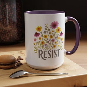 May include: White ceramic mug with a purple interior and handle. The mug features a floral design with yellow, pink, and purple flowers, and the word "RESIST" in gray lettering. The mug is on a wooden surface.