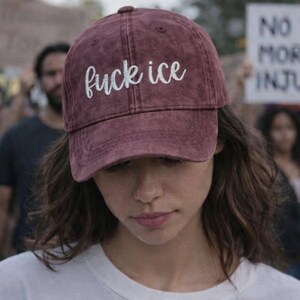 May include: A burgundy baseball cap with the text "fuck ice" embroidered in white cursive. The cap is worn by a person in a white t-shirt, with a blurred background of people and signs.