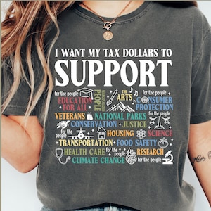 May include: A grey t-shirt with the text "I want my tax dollars to support" printed in white. The text is surrounded by a variety of colourful icons and text representing different causes, such as education, veterans, national parks, and climate change.