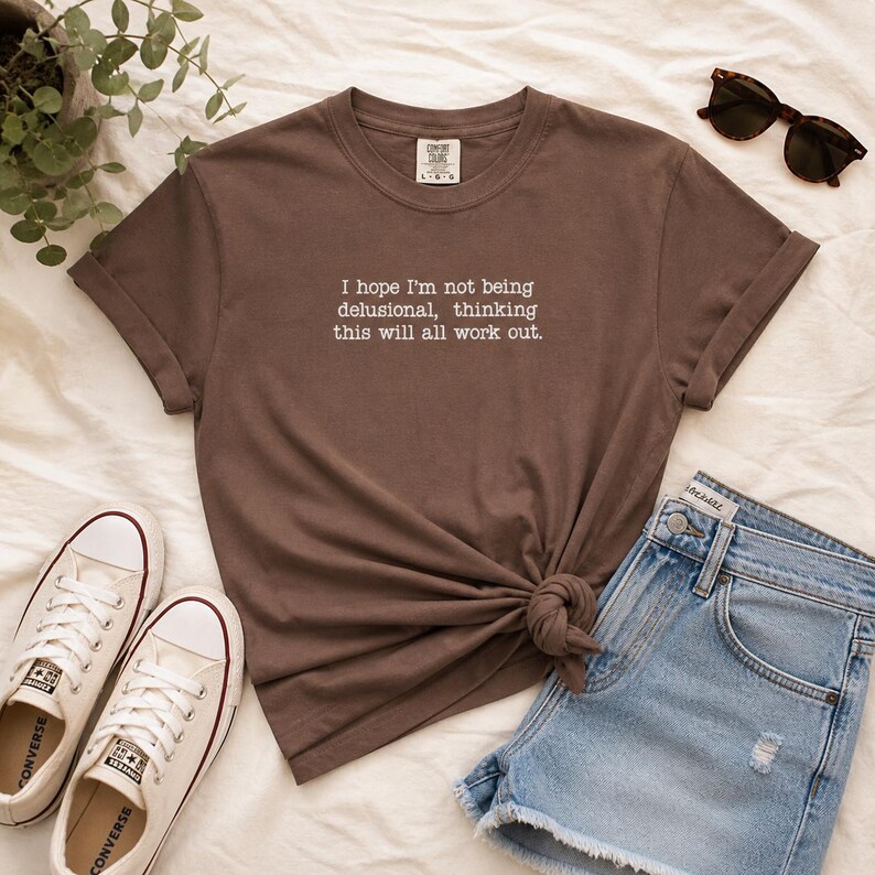 May include: A brown t-shirt with the text "I hope I'm not being delusional, thinking this will all work out." The shirt is styled with denim shorts, sunglasses, and white trainers.