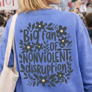 May include: A periwinkle blue sweatshirt with the text "Big fan of Nonviolent Disruption" in a floral design. The design features dark grey flowers and leaves. The sweatshirt is a casual style.