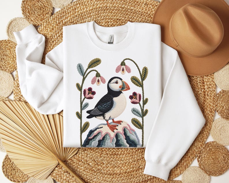 May include: White sweatshirt featuring a colorful embroidered puffin bird design. The puffin is perched on a rock with flowers and foliage. The design includes shades of black, white, blue, green, pink, and orange. The sweatshirt has long sleeves and a crew neck.