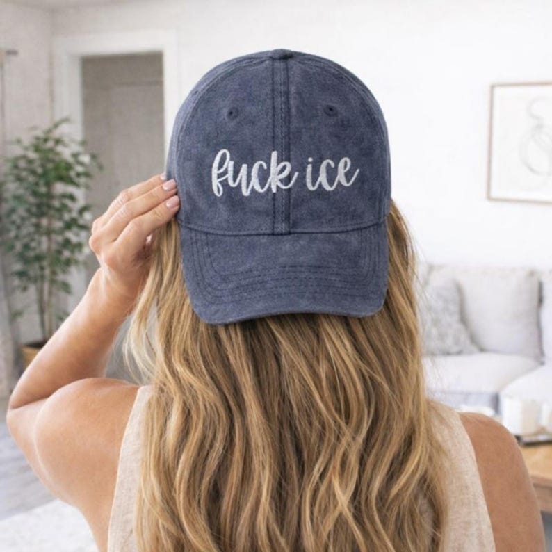 May include: A navy blue baseball cap with the phrase "fuck ice" embroidered in white script. The hat is worn by a person with long blonde hair, viewed from the back. The background is a softly lit interior.
