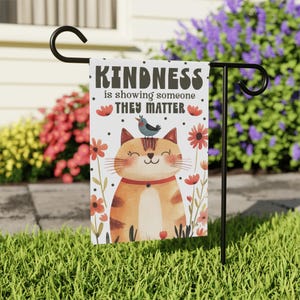 May include: A white garden flag with a cute orange cat and a blue bird on top of its head. The flag has black polka dots and red flowers. The text on the flag reads "Kindness is showing someone they matter."