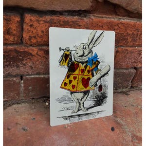 Puede incluir: Una tarjeta rectangular con una ilustración en blanco y negro del Conejo Blanco de Alicia en el País de las Maravillas. El conejo lleva un abrigo amarillo con corazones rojos y una pajarita azul, sosteniendo un pergamino y una trompeta. Fondo de pared de ladrillo.