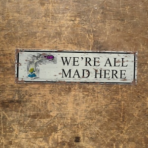 Könnte beinhalten: Ein weißes Metallschild mit schwarzem Text, der "We're All Mad Here" lautet. Das Schild zeigt ein Cartoon-Bild von Alice aus Alice im Wunderland.