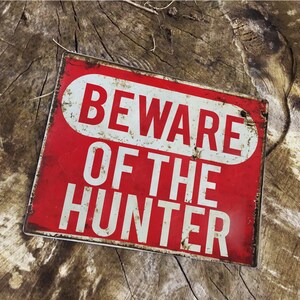 Puede incluir: Un letrero de metal rojo con texto blanco que dice "Beware of the Hunter". El letrero tiene un aspecto desgastado y vintage.