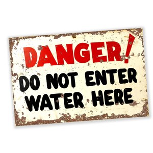Puede incluir: Un letrero de metal oxidado con un mensaje de advertencia en letras rojas y negras. El letrero dice "¡PELIGRO! NO ENTRAR AL AGUA AQUÍ."