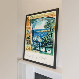 Puede incluir: Una impresión enmarcada de una pintura de Pablo Picasso que representa una vista de ventana de un paisaje marítimo con pájaros y ovejas. La pintura se titula "Côte d'Azur" y está firmada "Picasso".
