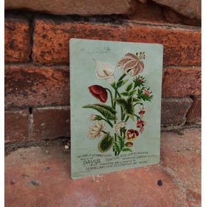 Puede incluir: Un anuncio vintage para la máquina de coser Davis Vertical Feed. El anuncio presenta un ramo de flores colorido, que incluye lirios, amapolas y pensamientos, sobre un fondo azul claro. El texto dice "Facsimile Embroidery Done on the Davis Vertical Feed Sewing Machine Pioneers and Leaders in Sewing Machine Decorative Art Work."