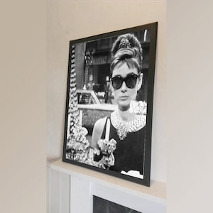 Puede incluir: Fotografía en blanco y negro de una mujer con gafas de sol y un collar de perlas. La mujer mira a la cámara y tiene el pelo recogido en un moño. La fotografía está enmarcada en un marco negro.