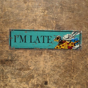 Puede incluir: Un letrero rectangular azul turquesa con el texto "I'M LATE" en letras negras. El letrero presenta una imagen de dibujos animados del Conejo Blanco de Alicia en el País de las Maravillas con un traje rojo y amarillo con corazones y una pajarita azul. Sostiene una trompeta en su mano derecha.