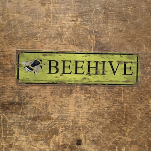 Puede incluir: Un letrero de metal amarillo y verde con borde negro y la palabra "BEEHIVE" en letras negras. Una abeja negra y amarilla está representada en el lado izquierdo del letrero.