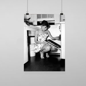 Puede incluir: Una mujer con un vestido floral está agachada en una cocina, mirando dentro de un horno abierto. Está sosteniendo una bandeja para hornear en sus manos.