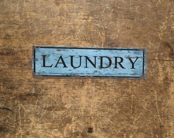 May include: A rectangular, distressed blue sign with the word "LAUNDRY" in black capital letters. The sign has a rustic, vintage aesthetic, with visible wear and tear, and is set against a textured wooden background.