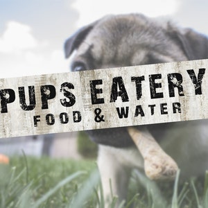 Puede incluir: Un letrero de madera blanco con texto negro que dice "Pups Eatery Food & Water". Un perro carlino está en primer plano, oscureciendo parcialmente el letrero.