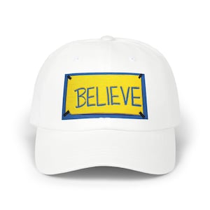 Puede incluir: Una gorra de béisbol blanca con un parche rectangular en la parte frontal. El parche tiene un fondo amarillo con un borde azul y detalles negros en las esquinas. La palabra BELIEVE está bordada en letras mayúsculas azules en el centro.