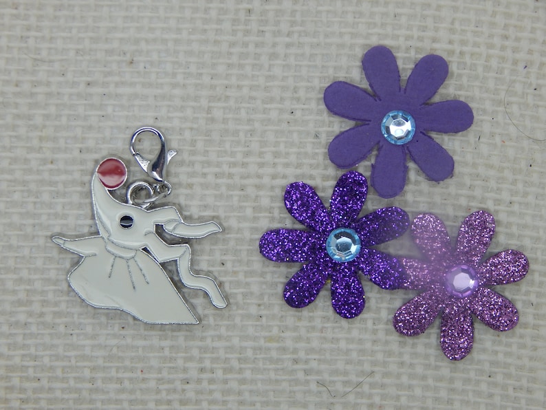 May include: A white metal charm of the character Zero from the movie The Nightmare Before Christmas. Zero is holding a red ball in his mouth and is wearing a white dress. The charm is attached to a silver metal ring. The charm is surrounded by three purple glitter flowers with blue glitter centers.