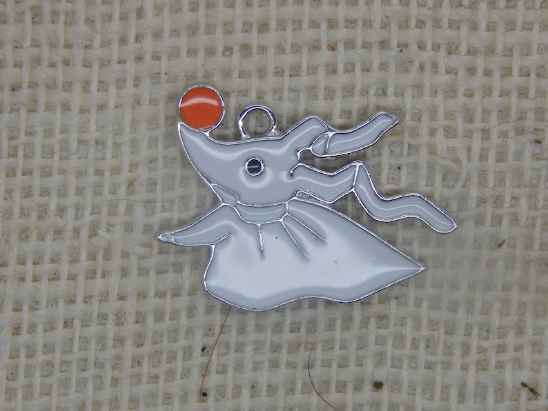 May include: White metal charm of a ghost character with an orange ball on top of its head. The ghost has a long, flowing tail and a single eye.