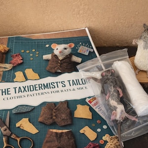 May include: A book titled "The Taxidermist's Tailor" with clothes patterns for rats and mice. The image includes a stuffed mouse wearing a vest, sewing supplies, and a taxidermied rat. A second taxidermied rat stands on a wooden disc.