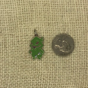 May include: Green Grinch character charm with a red heart and a blue flower. The charm is made of metal and has a shiny finish. The charm is about the size of a US quarter.
