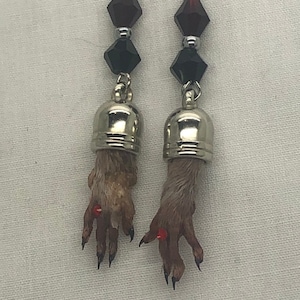 May include: A pair of unique earrings featuring small, taxidermied animal feet. Each earring has a silver-colored cap, black and red faceted beads, and a silver hook. The animal feet are brown with visible claws.